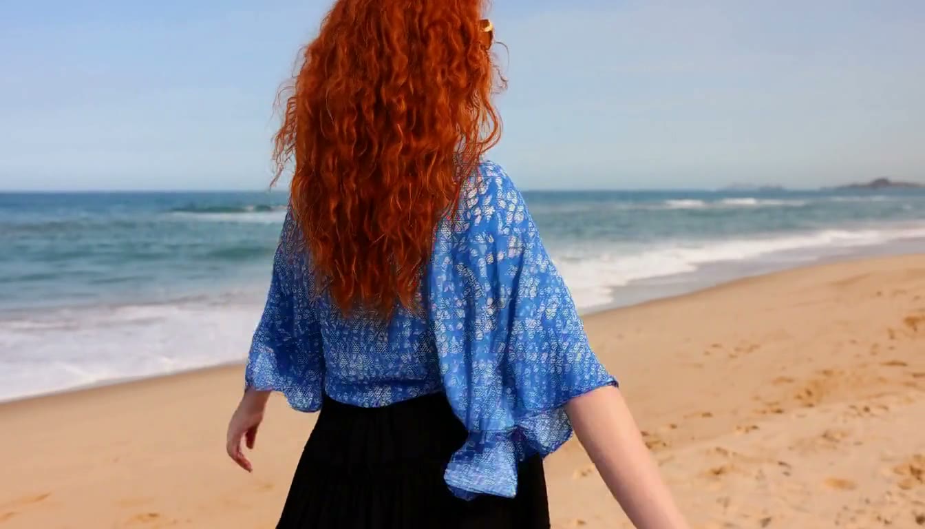 MiniMax H3 AI video example showing a woman with curly red hair walks along a sandy beach, wearing a blue floral top and a black skirt, with the ocean waves in the background