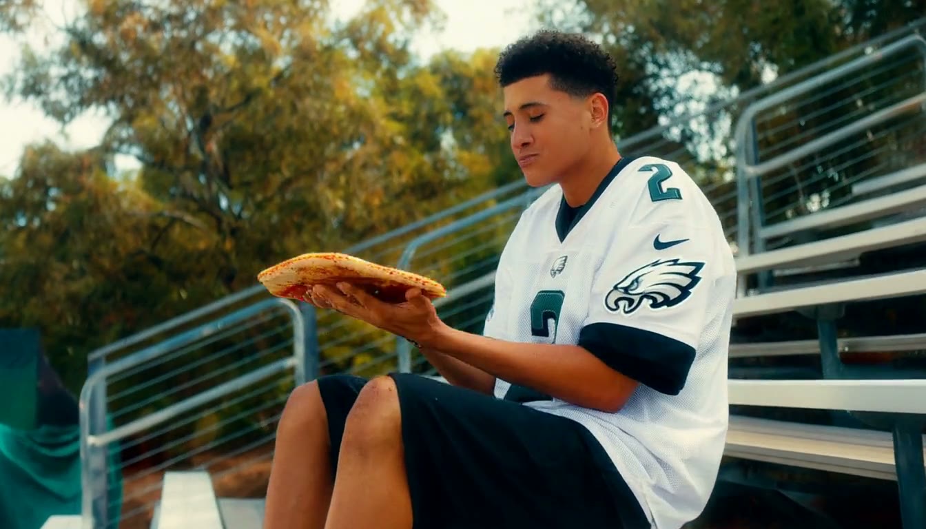 MiniMax H3 AI video example showing a young man in an sports uniform is sitting on bleachers surrounded by pizza, enjoying a playful moment